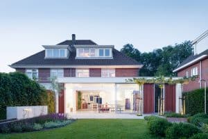 Solarlux zeigt ein rotes Einfamilienhaus mit Dachschrägen und Glasfaltwand, im Garten ist viel Grünfläche und eine Outdoordusche.