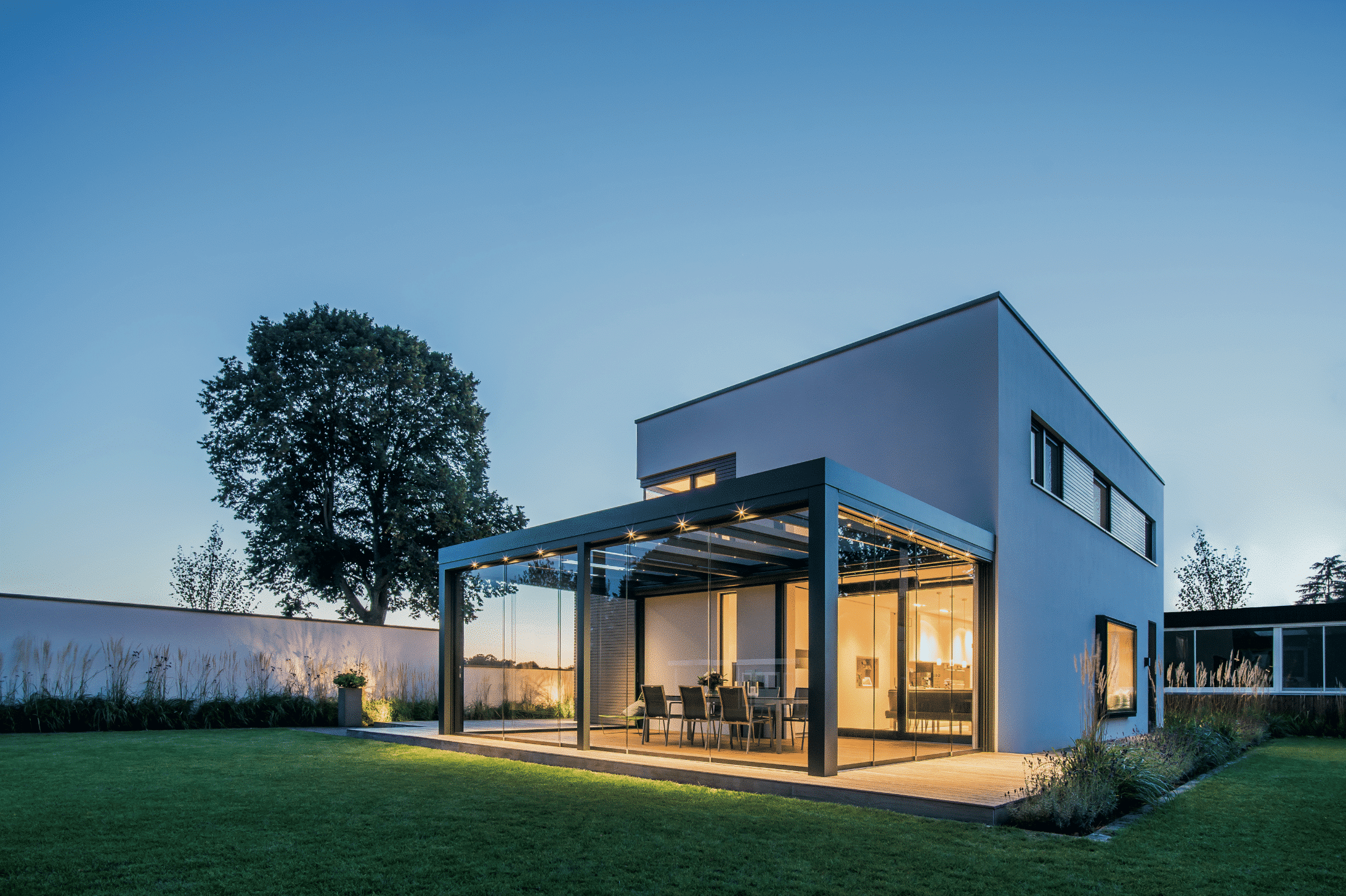 Solarlux zeigt ein Einfamilienhaus mit barrierefreiem Wintergarten in dem Outdoormöbel stehen und man direkt über eine kleine Stufe in den Garten kann.