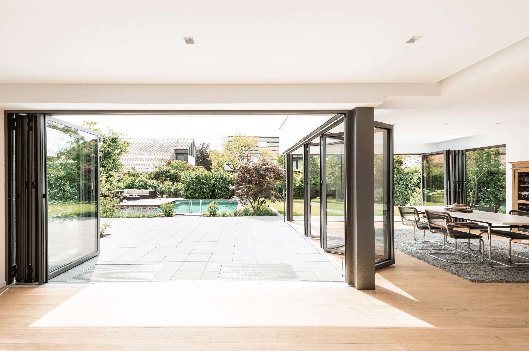 Solarlux zeigt ein offenes Esszimmer mit Holzboden, direktem Zugang zur gefliesten Terrasse durch eine Glasfaltwand und Weg in den Garten mit Pool.