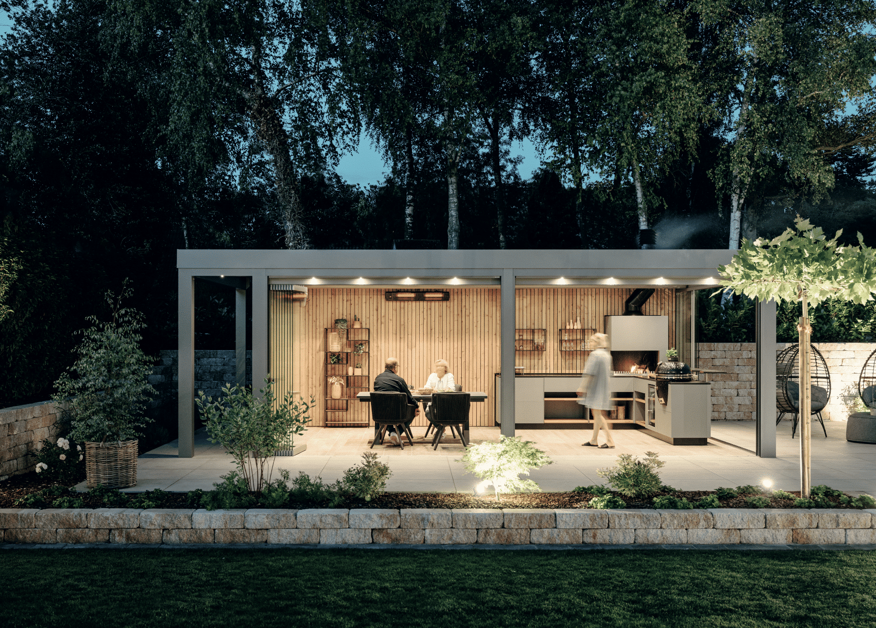 Solarlux zeigt einen Garten mit kleiner Ziegelmauer und überdachter Terrasse mit Holzverkleidung an der Rückwand, Beleuchtung und Verglasung, mit einer Outdoorküche und gemütlicher Sitzgruppe.