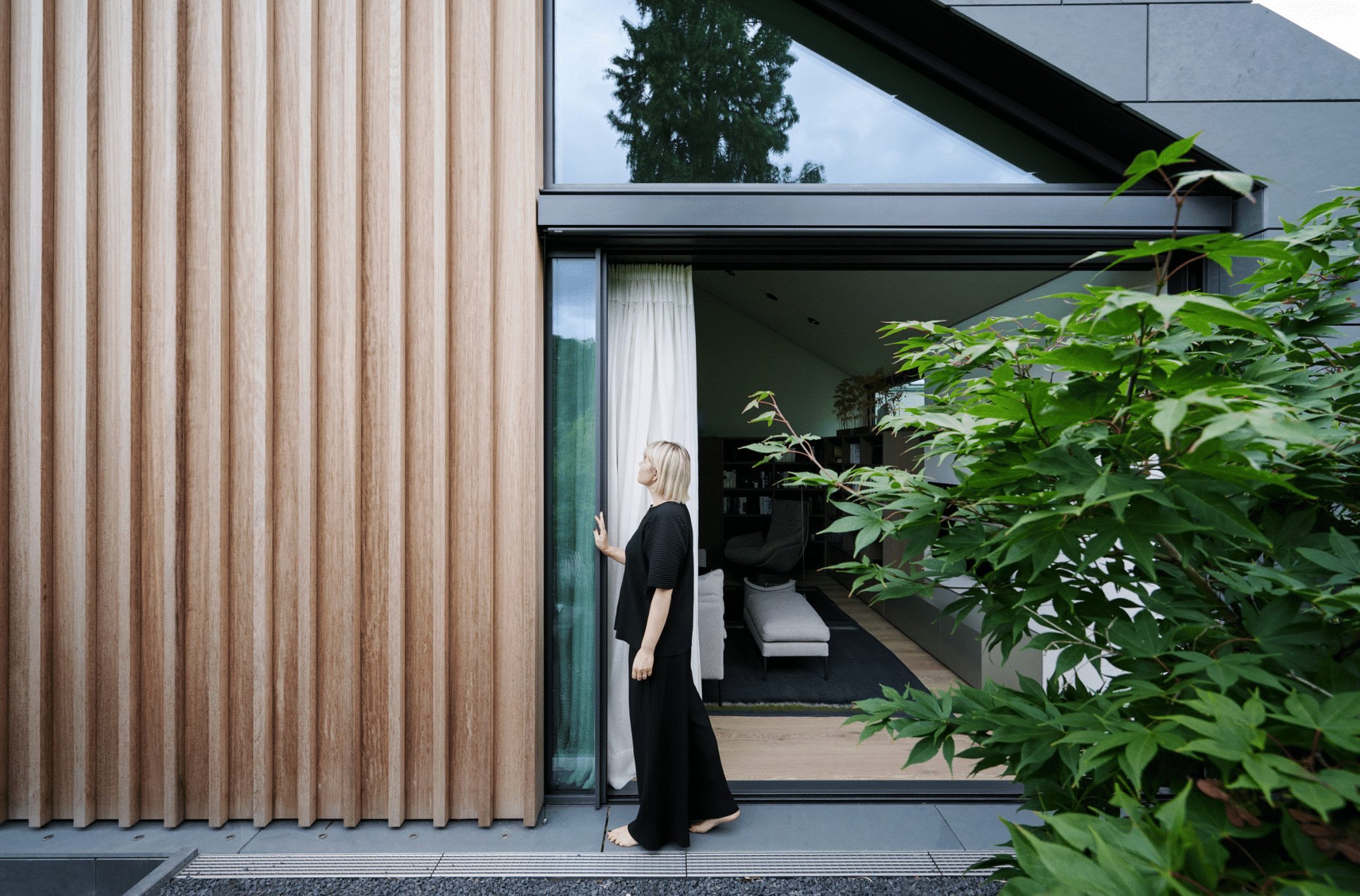 Solarlux zeigt den Außenbereich eine Hauses mit Holzverkleidung und Fliesenboden und dem Zugang von der Terrasse in das Wohnzimmer durch eine große Glasschiebetür.