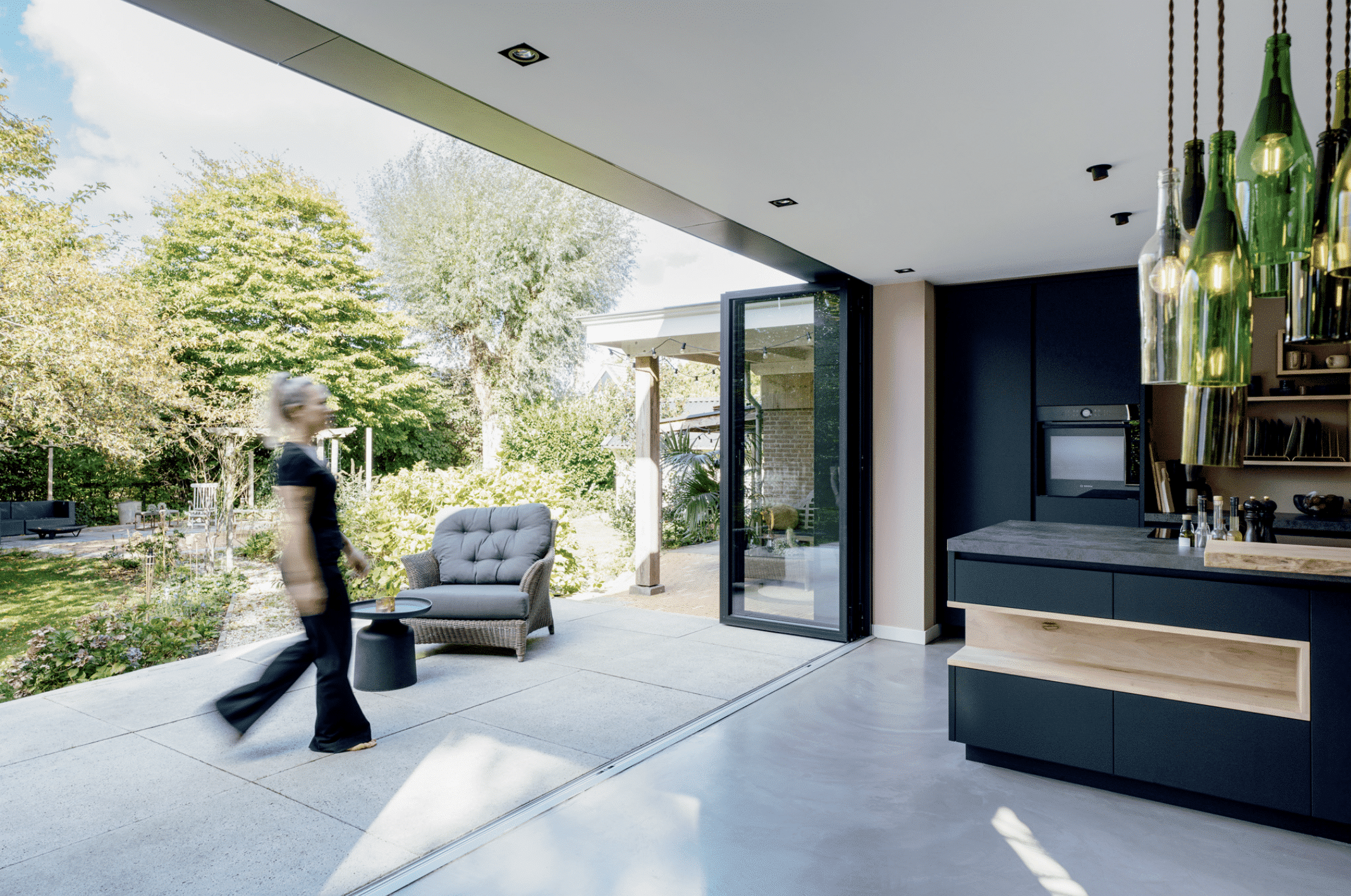 SOLARLUX präsentiert ein Glasfaltfenster, welches von der schwarzen Küche mit Holzelementen auf die Terrasse mit Outdoormöbeln führt.