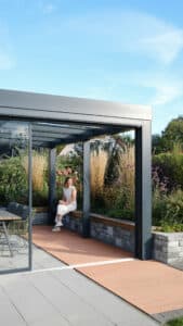 Ein modernes Glashaus „Acubis“ mit anthrazitfarbenem Rahmen, Glasdach, Glasschiebetüren integriert in eine stilvolle Aussenanlagengestaltung mit Ziegelmauer und Sitzgelegenheit von Solarlux.