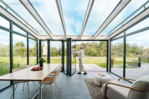 Ein moderner Wintergarten „SDL Avantgarde“ mit Esstisch und Loungebereich wo eine Frau die schwarzgerahmten Glasfalttüren zum Garten öffnet - von Solarlux.