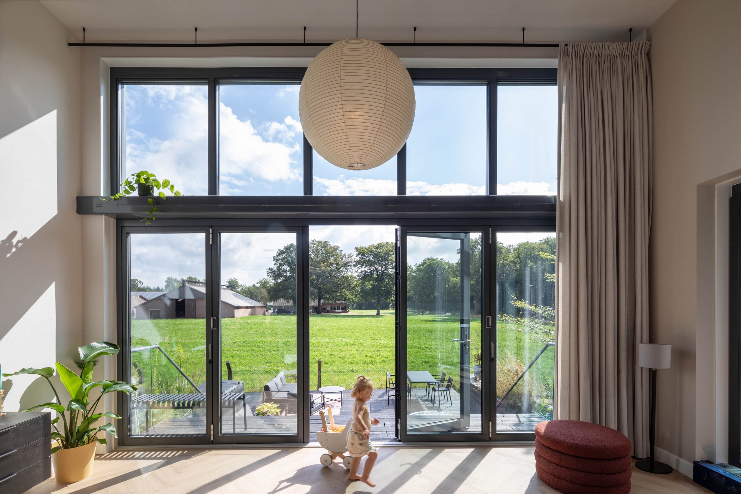 Ein kleines Mädchen spielt in einem Wohnzimmer mit Glasfalttüren mit Oberlichte und Rahmen in Schwarz von Solarlux. Es sind 5 Glaselemente wobei die Tür in der Mitte geöffnet ist - links befindet sich die 2-flügelige und rechts die 3-flügelige Glasfalttüre von Solarlux.