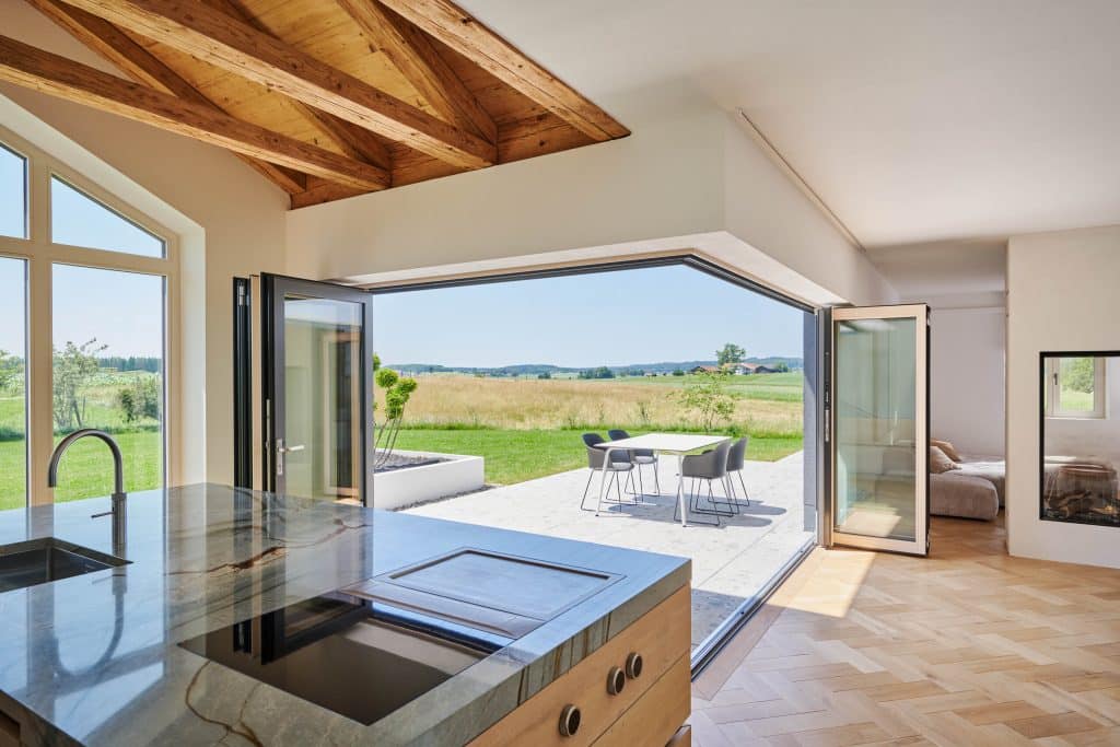 Eine offene Wohnküche mit Erker und Sichtdachstuhl und über das Eck vollkommen geöffneten Glasfaltwänden zur Terrasse mit Essbereich von Solarlux.