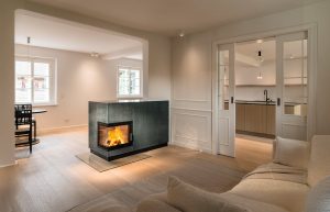 Sommerhuber zeigt modernen rechteckigen Kachelofen aus grün strukturieren Kacheln mit Ecksichtfenster als Raumtrenner in einem offen Wohnzimmer von Sommerhuber.