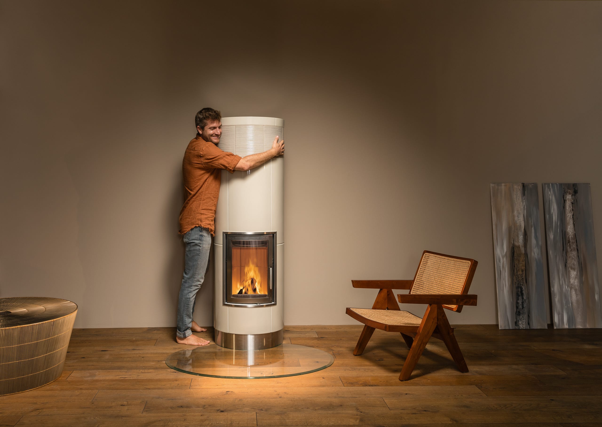 Sommerhuber zeigt eleganten säulenförmigen Vollspeicherkachelofen aus beige strukturieren Kacheln mit silber umrandeter Ofentüre, silber Sockel auf runder Glasscheibe von Sommerhuber.