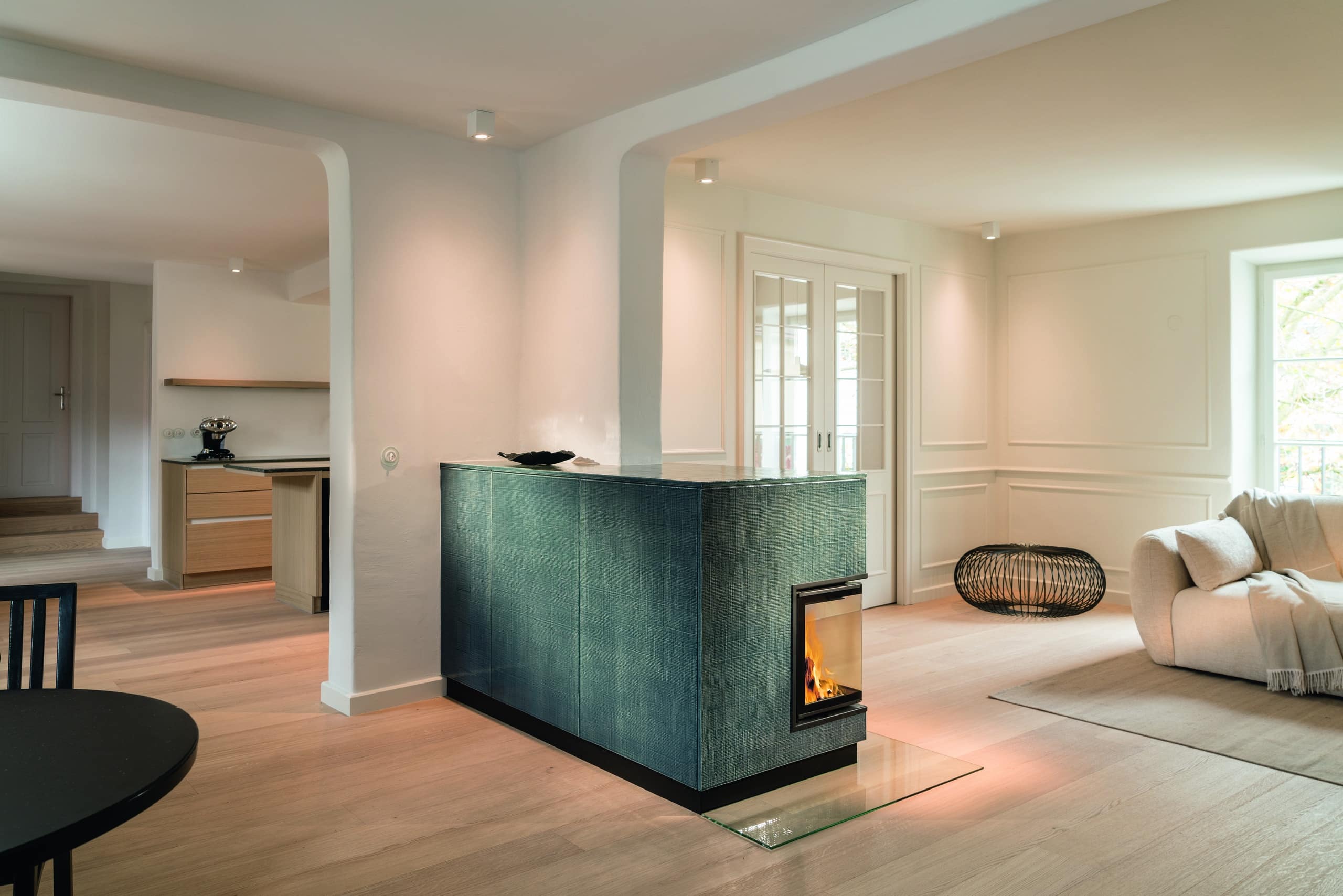 Sommerhuber zeigt modernen rechteckigen Kachelofen aus grün strukturieren Kacheln mit Ecksichtfenster als Raumtrenner in einem offen Wohnzimmer von Sommerhuber.