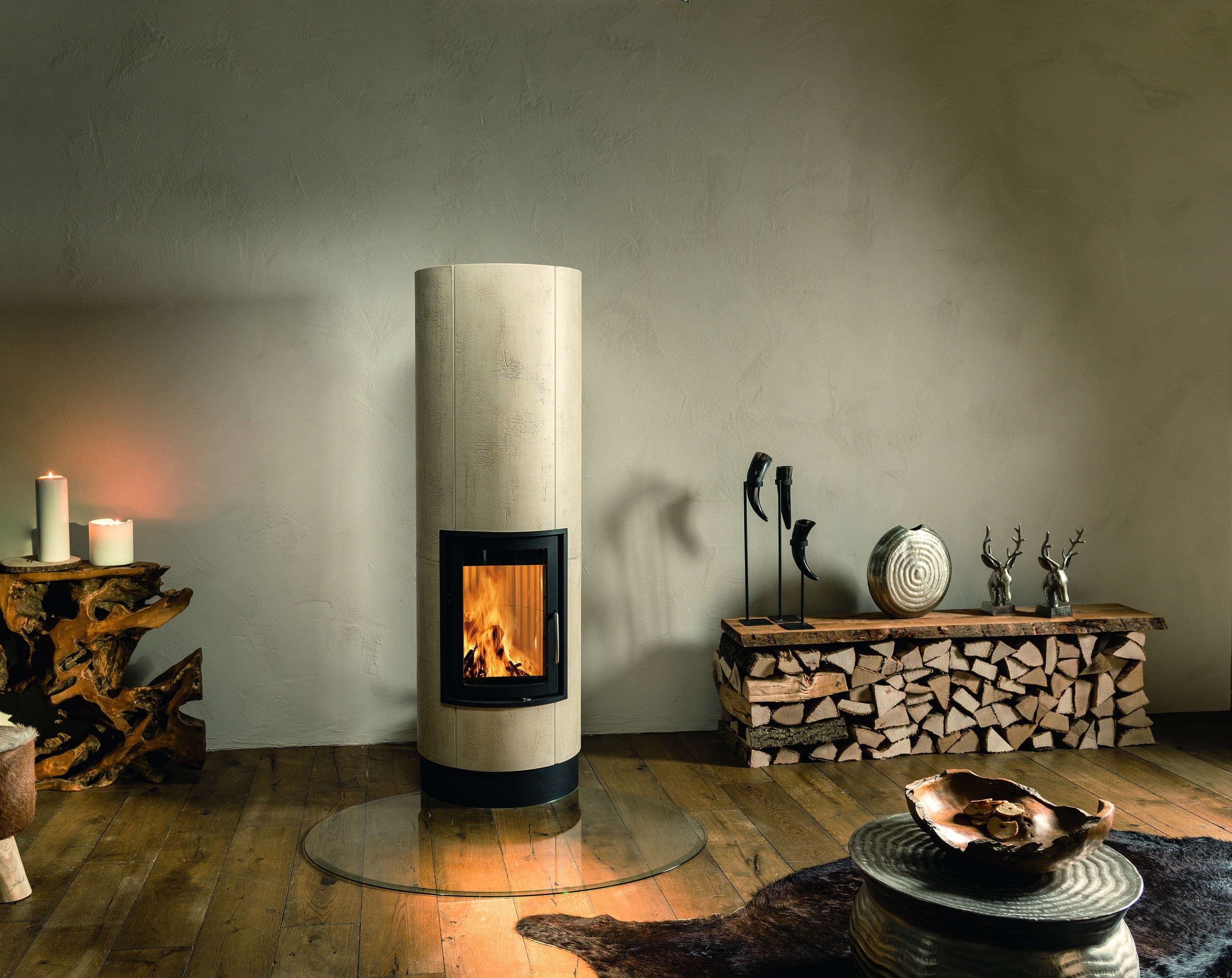 Sommerhuber zeigt eleganten säulenförmigen Vollspeicherkachelofen aus beige strukturieren Kacheln mit schwarz umrandeter Ofentüre, schwarzem Sockel auf runder Glasscheibe von Sommerhuber.