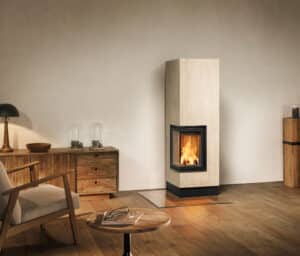 Ein hoher kubisch-quadratischer Kamin mit beige-warmweißen rechteckigen Keramikfliesen und Ecksichtfenster mit schwarzem Rahmen eingebettet in eine rustikale Einrichtung bestehend aus warmen Hölzern von Sommerhuber.