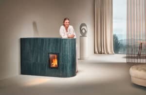 Eine Dame lehnt entspannt an einem Kamin mit abgerundeten Aussenkanten, Relief-Keramik in Stäbchen-Optik in einem Blau bzw. Petrolton und Kamintüre mit Sichtfenster von Sommerhuber.