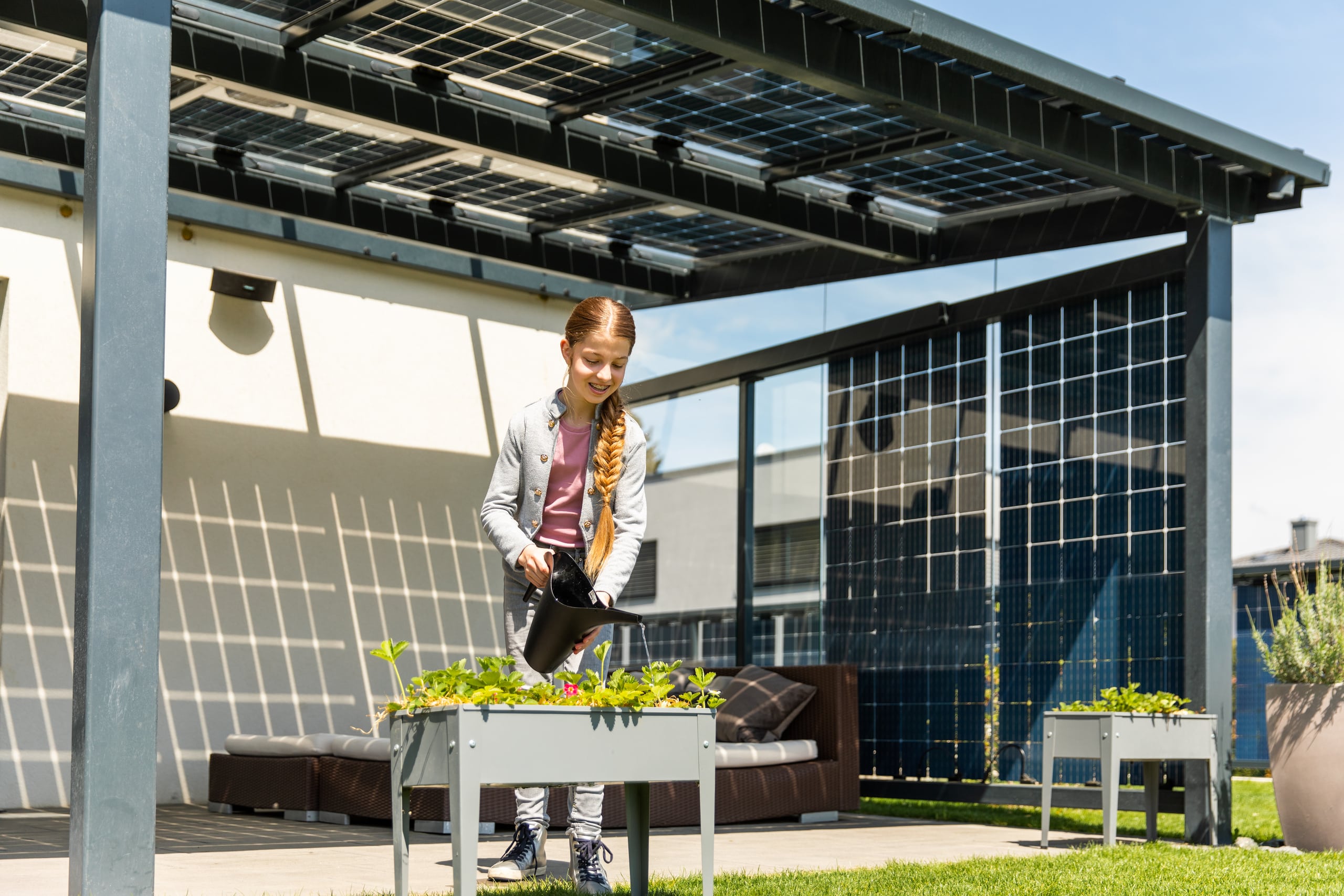 Ein kleines Mädchen gießt ein Pflanzbeet unter einer Glaspergola, am Dach und an den Seiten gespickt mit vielen quadratischen Solar-Modulen von Sonnenkraft.