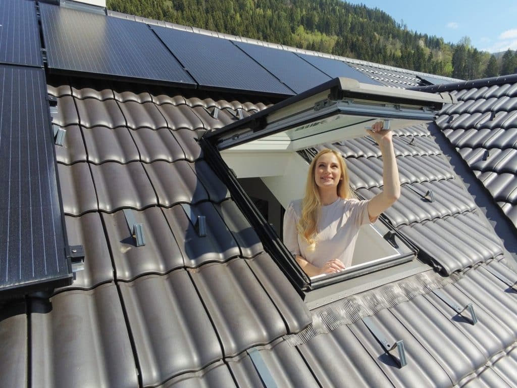 Ein junge Frau öffnet ein Dachfenster und betrachtet den blauen Himmel und ihre vielen großformatigen dachmontierten Solar-Module von Sonnenkraft.