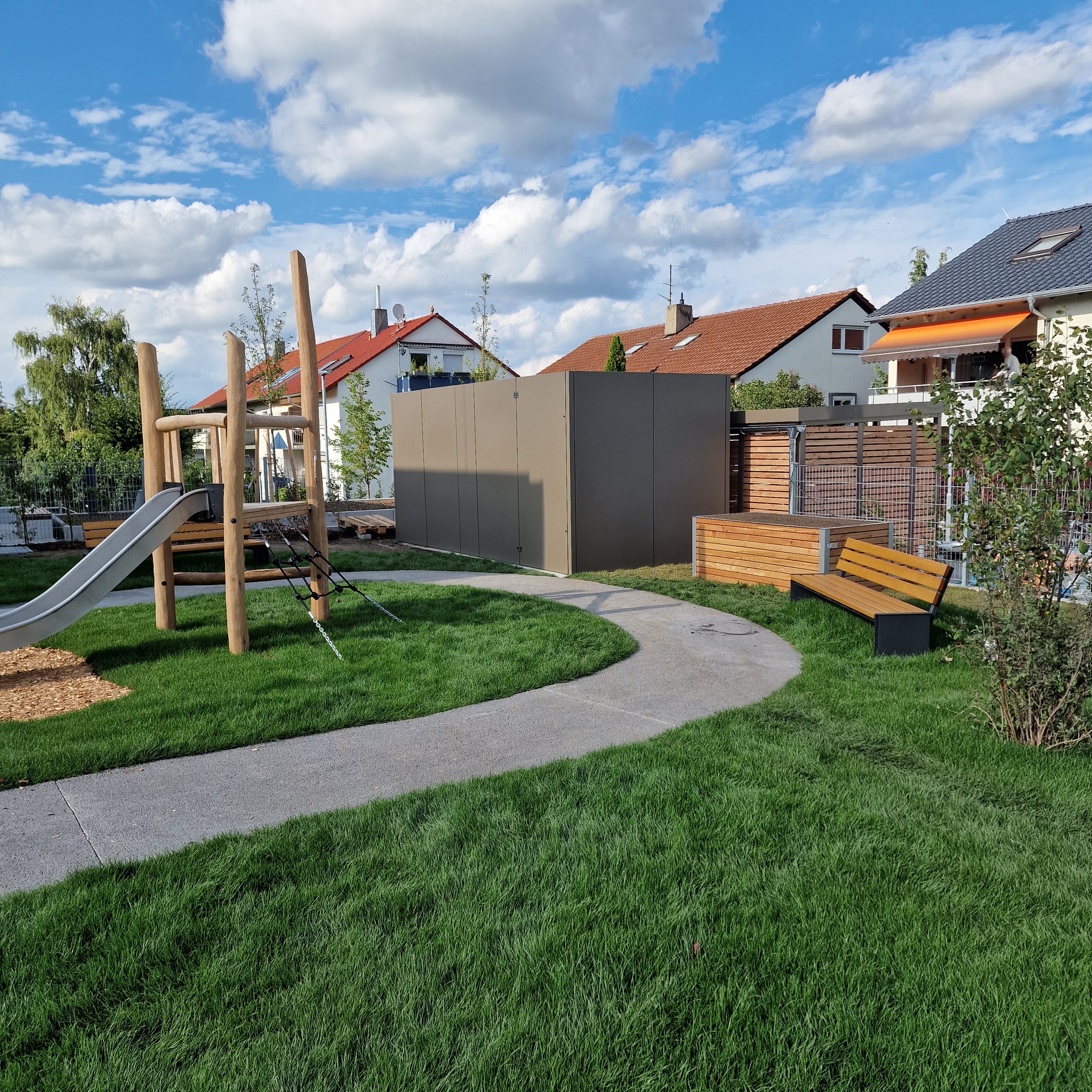H Stocker zeigt den Spielplatz eines Kindergartens in einer Wohnsiedlung, auf dem eine große Wärmepumpe mit dunkelgrauem Schallschutzgehäuse steht.
