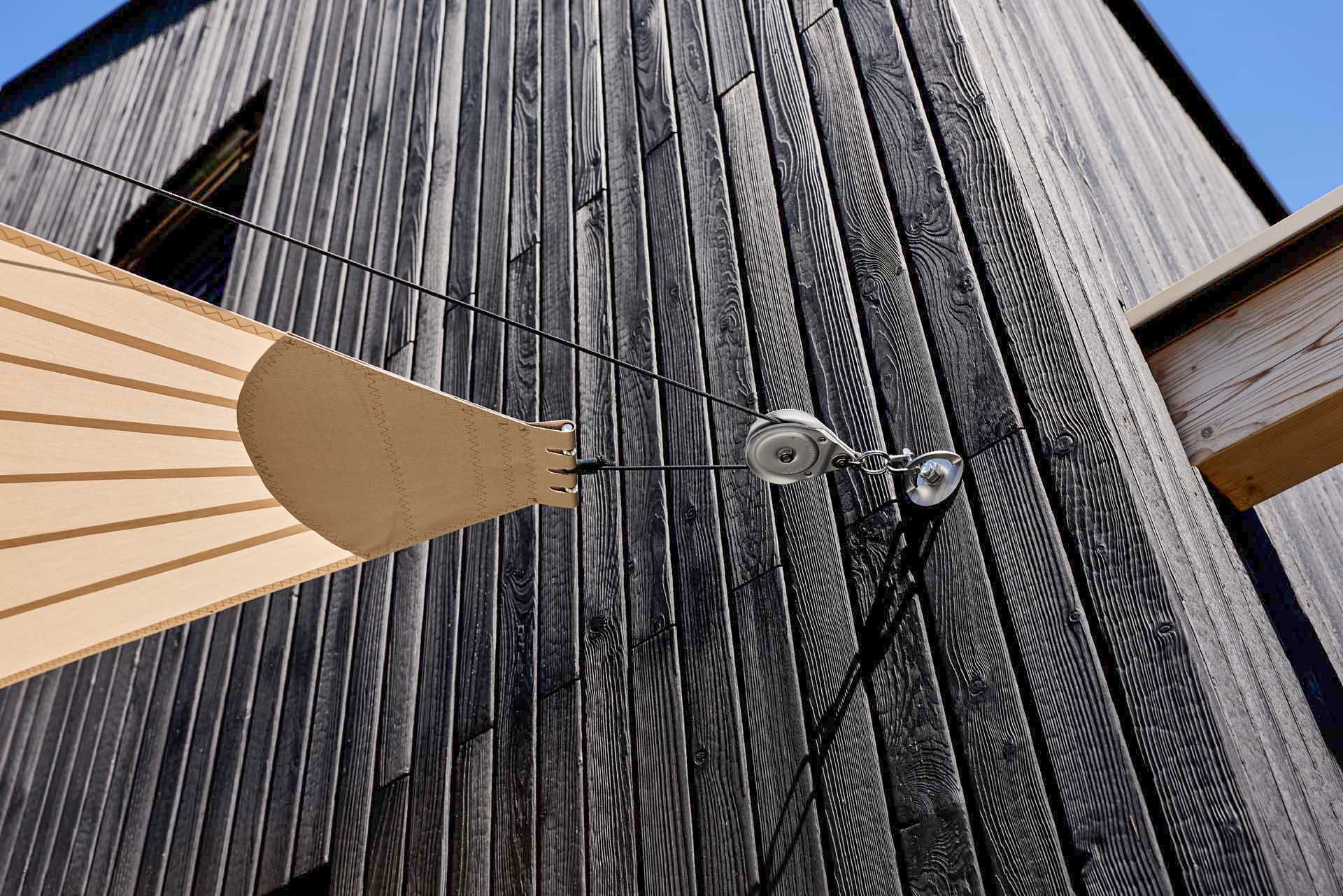 SunSquare Shading Solutions zeigt die Befestigung eines Sonnensegels mittels Umlenkrolle an der Holzfassade eines Einfamilienhauses.