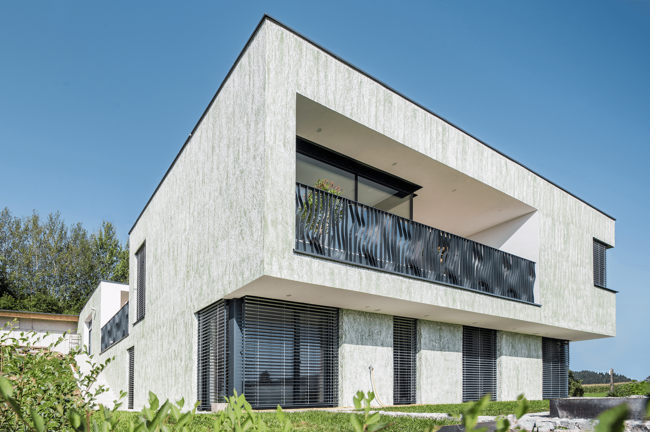 SynthesaGruppe zeigt ein Einfamilienhaus mit Flachdach, grauer Fassade, einem Balkon mit Metallgeländer in geschwungener Optik und einer Terrasse.