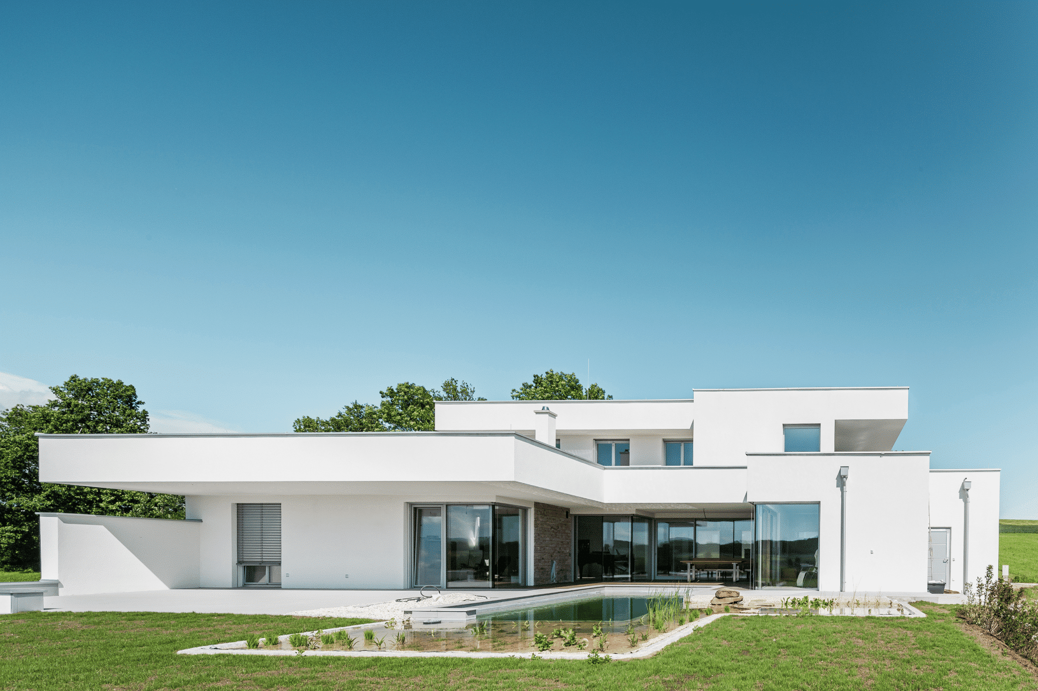 Modernes weisses Einfamilienhaus mit Flachdach, grossem Garten mit Pool und einer überdachten gefliesten Terrasse von SynthesaGruppe.