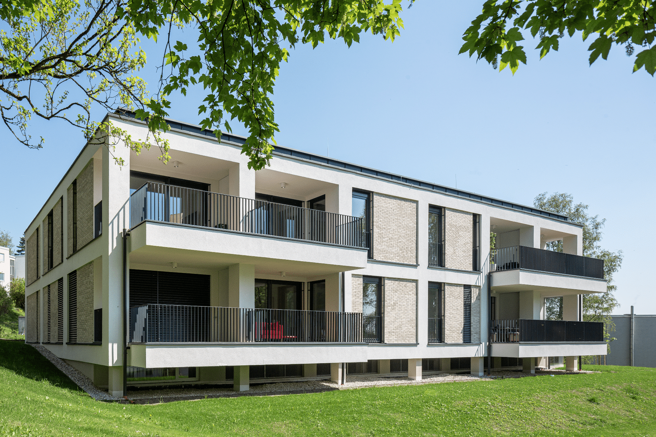 SynthesaGruppe zeigt ein Mehrfamilienhaus im Grünen mit Flachdach und Balkon mit schwarzem Geländer bei jeder Wohnung.