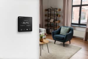 Technische Alternative RT zeigt ein Raumthermostat an der Wand eines Wohnraumes montiert, mit mattem Display das die Temperatur anzeigt.