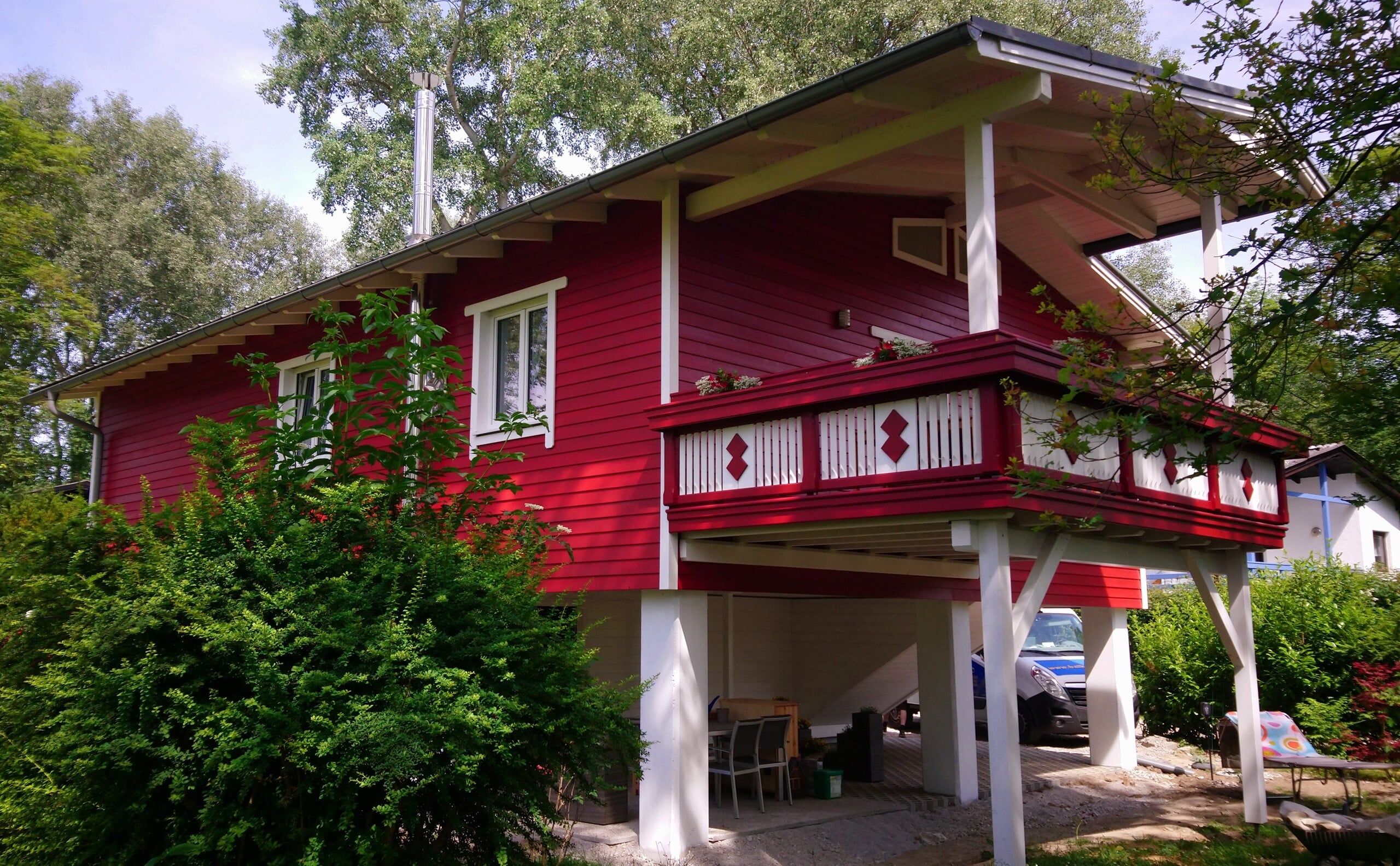 Ferienhaus mit Fassadenverkleidung aus Holz und großzügigem überdachtem Balkon mit verspielten Holzelementen am Geländer in Rot und Weiss lackiert von Richrad Teufel.