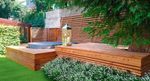Gartenbereich mit erhöhtem Holzpodest und Aussparungen für Aufstellpool oder Whirlpool und einem schattenspendenden Baum und einer an einer Betonmauer entlang verlaufenden schrägen Holzsprossenwand die auch als Pflanzwand oder Kletterhilfe dienen kann von Richard Teufel.