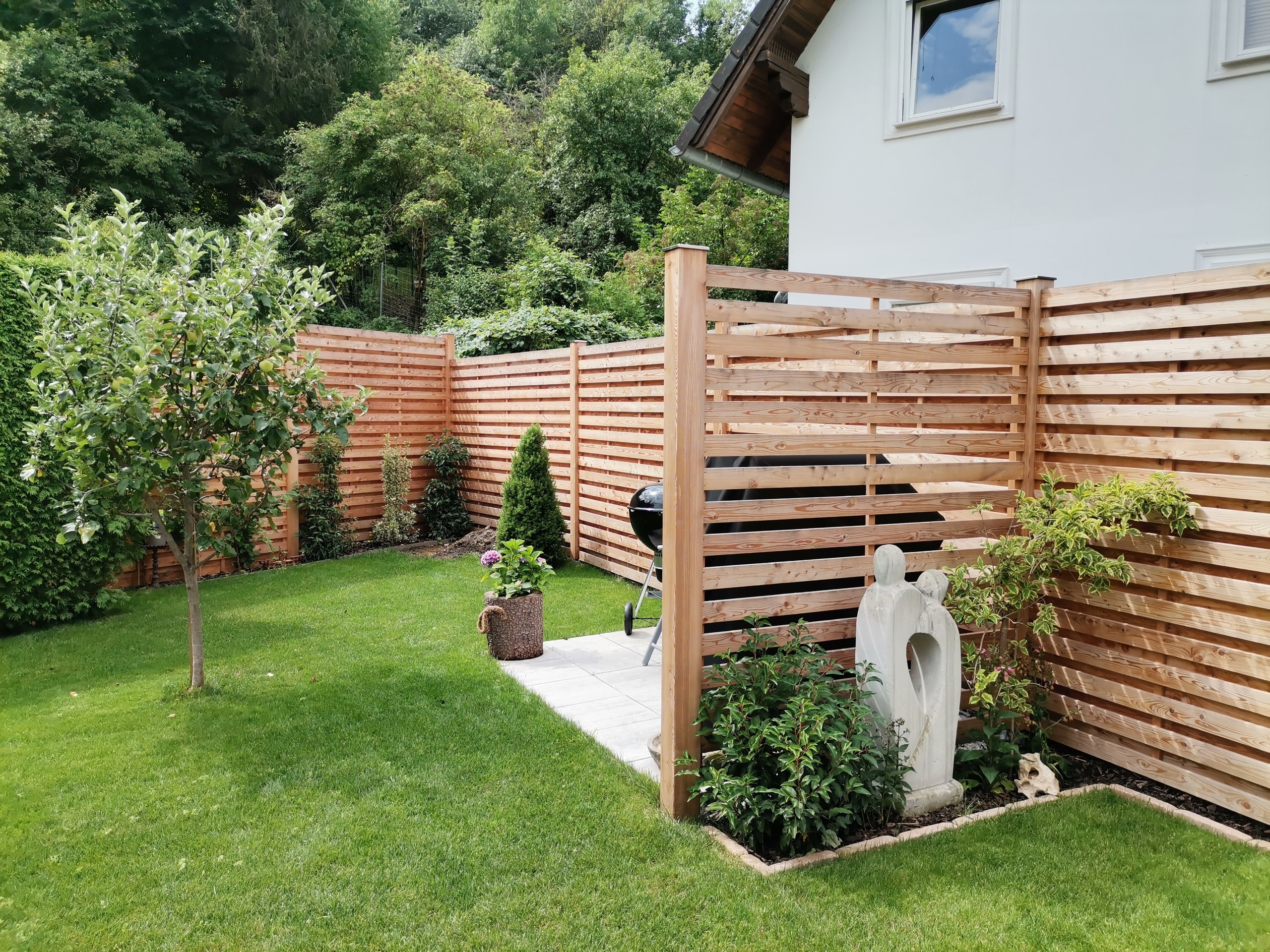 Garten mit hohem Gartenzaun aus versetzten Holzpaneelen mit horizontalen Sprossen von Richard Teufel die sowohl praktisch als auch funktional als Abtrennung für Grillbereich und Beete dienen.