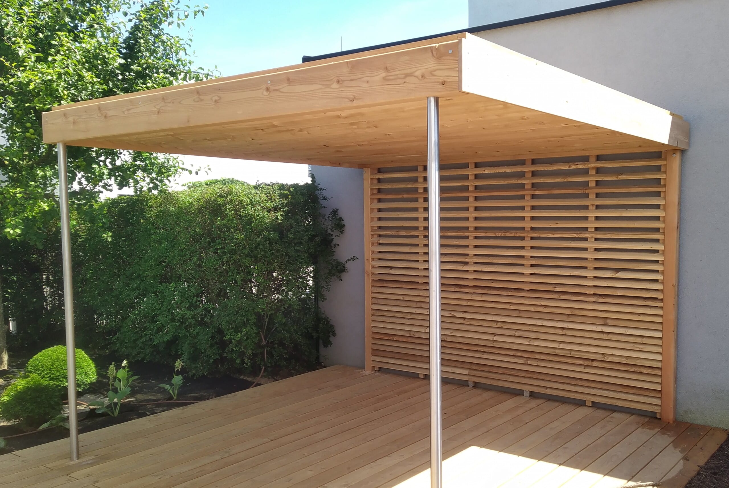 Offene, moderne Pergola in hellem Holz mit Holzdielen, einer Rückwand mit waagrechten Sprossen und runden Eisenstäben als Träger von Richard Teufel.