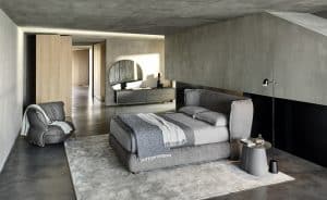 Bauer Tischlerei zeigt Schlafzimmer mit Boxspringbett mit hohem Kopfteil und grauem Stoffbezug, Beistelltisch und grauen Sessel mit dicker Polsterung von Ditre Italia.