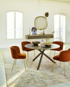 Bauer Tischlerei zeigt runden Dreibein-Esstisch aus dunklem Holz, Stühle mit orangem Stoffbezug und goldenen Beinen und gold-braunes Sideboard von haas Möbelwerk.