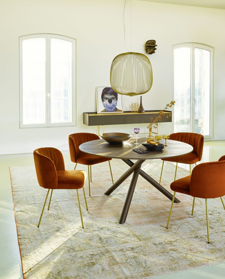 Bauer Tischlerei zeigt runden Dreibein-Esstisch aus dunklem Holz, Stühle mit orangem Stoffbezug und goldenen Beinen und gold-braunes Sideboard von haas Möbelwerk.