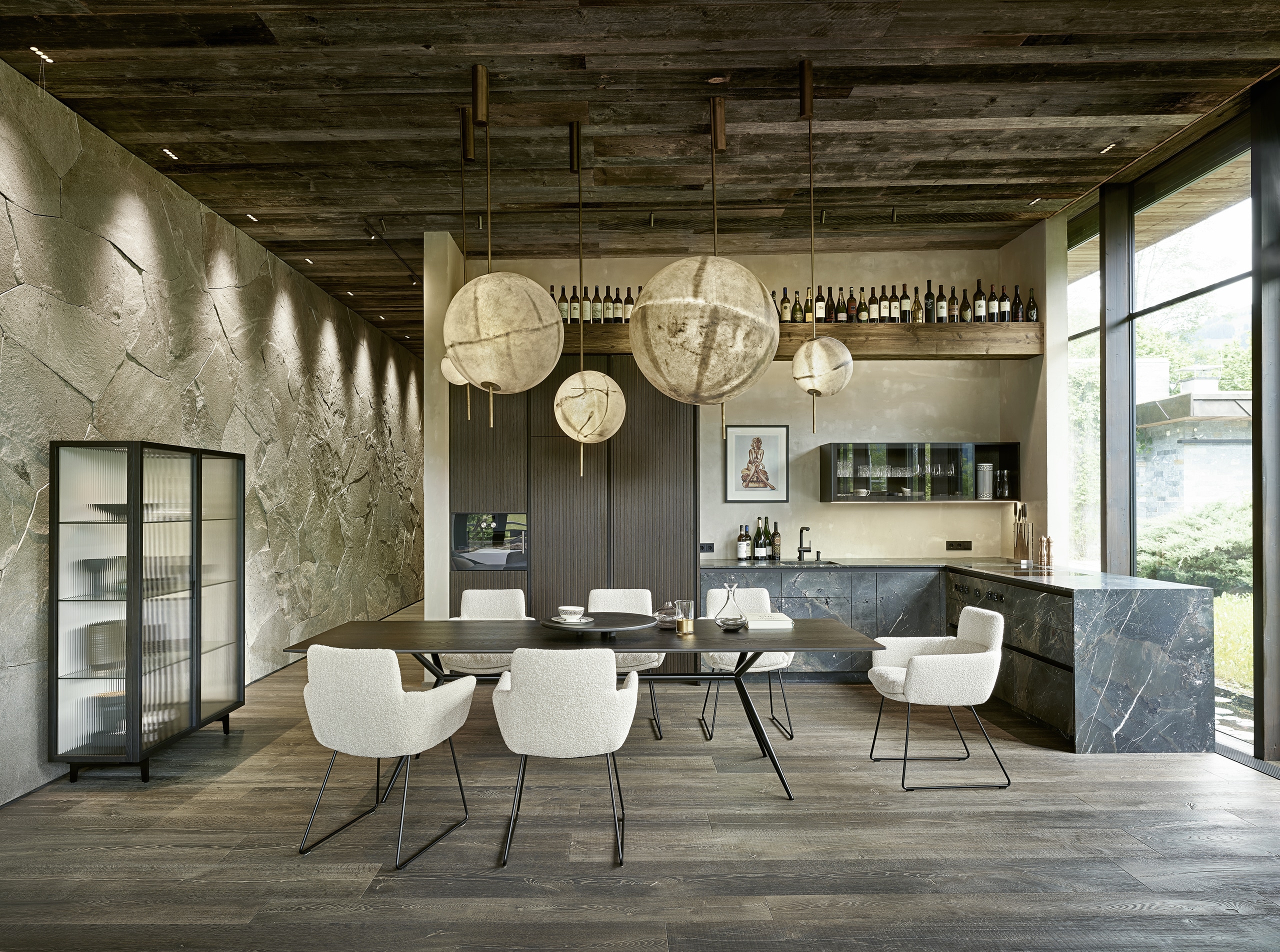 Bauer Tischlerei zeigt Küche mit grifflosen Mamorfronten, Hochschrank mit Holzfront, Hängeschrank mit Glasfront, zierlichen Esstisch mit Holzfläche und Metallbeinen und weiße Stoff-Stühle mit Armlehnen von haas Möbelwerk.