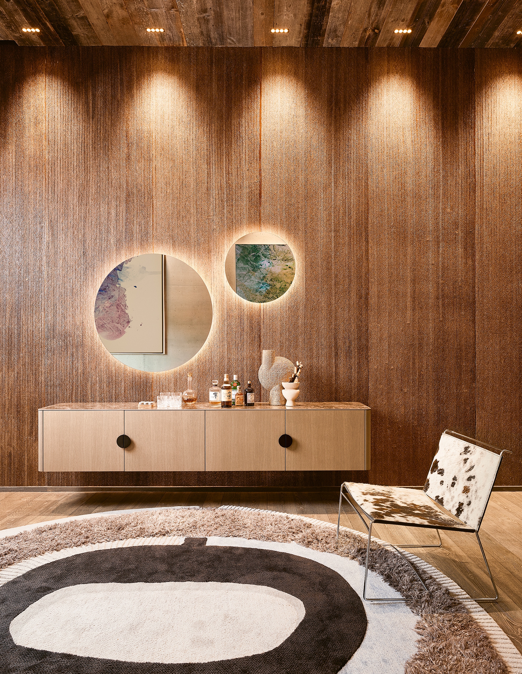 Ein rustikal stilistischer Vorraum mit retro-brauner Textiltapete, schwebender Holz-Garderobenkommode, rundem Spiegel, verspieltem freiliegendem Teppich in Brauntönen und Sessel in Kuhfelloptik von BAUER Tischlerei zeigt haas Möbelwerke.