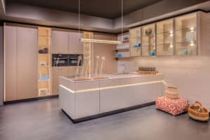 Ein heller, offener Küchenraum mit beleuchteten Eingriffmulden, offenen Regalen, glatten beigen Fronten, integrierten Geräten und einer Hängevitrine aus Glas mit Holzrahmen von BAUER Tischlerei zeigt Sachsenküchen.