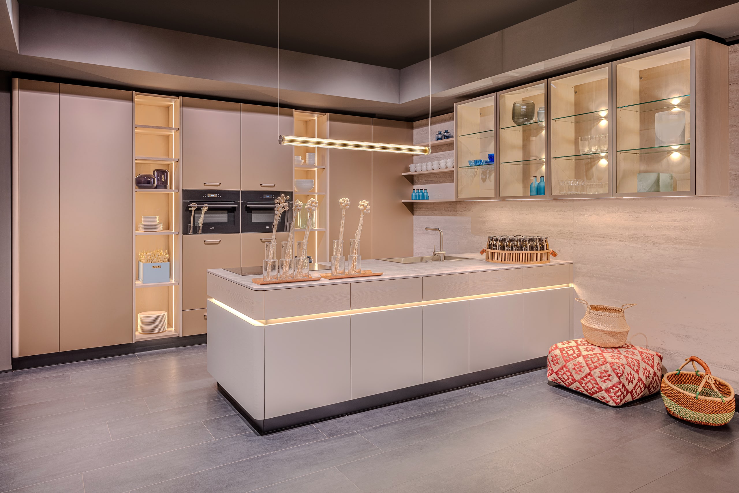 Ein heller, offener Küchenraum mit beleuchteten Eingriffmulden, offenen Regalen, glatten beigen Fronten, integrierten Geräten und einer Hängevitrine aus Glas mit Holzrahmen von BAUER Tischlerei zeigt Sachsenküchen.