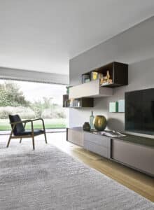 Ein modernes, stilvolles Wohnzimmer in Grautönen mit freiliegendem Teppich, Lounge-Chair, einem eleganten Medien-Board und filigranen Wandboards mit Materialmix von BAUER Tischlerei zeigt Sangiacomo.