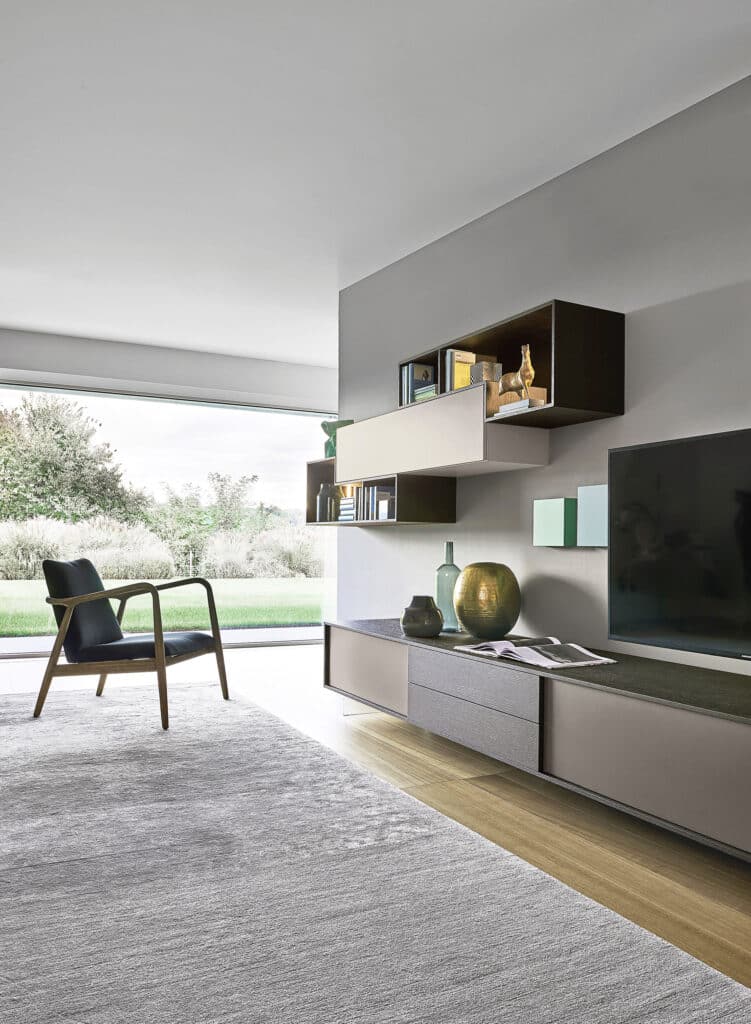 Ein modernes, stilvolles Wohnzimmer in Grautönen mit freiliegendem Teppich, Lounge-Chair, einem eleganten Medien-Board und filigranen Wandboards mit Materialmix von BAUER Tischlerei zeigt Sangiacomo.