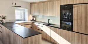 Deckenhohe Holzküche in U-Form mit Induktionsfeld mit Downdraft-Dunstabzug einer Arbeitsplatte in Schwarz und einer Küchenrückwand aus Glas von Tischlerei Penninger.