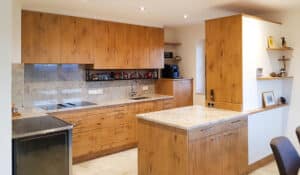 Eine u-förmige Küche mit deckenhoher Küchenzeile, grifflosen Fronten Holz mit Verästelungen, einer Arbeitsplatte und Küchenrückwand aus Stein und einer raumtrennenden Wandnische mit passender Theke von Tischlerei Penninger.