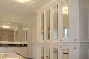 Verspieltes, luxuriöses Badezimmer in Weiss mit deckenhohen Kästen mit Kassettenfronten, eingearbeiteten Spiegeln und Goldknöpfen von Tischlerei Penninger.
