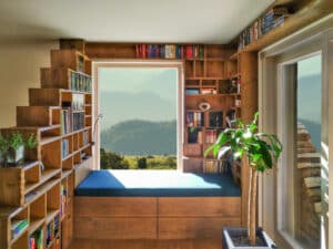 Eine massivverbaute, deckenhohe Vollholz-Leseecke mit einem Bücherregal, welches gleichzeitig als Raumteiler dient, einem integriertem, gepolsterten Sitzfenster mit Stauraum daneben eine Terrassenschiebetüre von Tischlerei Penninger.