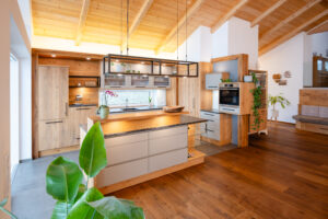 Tischlerei Scharnreithner zeigt eine Küche mit Holzfronten mit Holzmaserung und einer Kücheninsel mit dunkelgrauer Stein-Arbeitsplatte, Barfunktion und viel Stauraum.