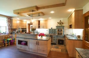 Tischlerei Scharnreithner präsentiert eine rustikale Küche mit Holzfronten und silber Griffen, einem Kamin mit Kochfeld und Backofen, dunkle Naturstein-Arbeitsplatte und einer Kochinsel mit erhöhter Ablagefläche.