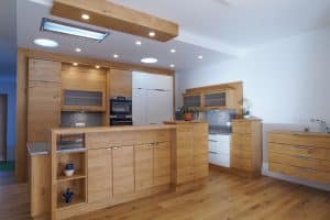 Tischlerei Scharnreithner zeigt eine Küche mit Holzfronten und weißen Fronten, Hängeschränke mit Milchglas, Arbeitsplatte und Rückwand in Steinoptik und eine Kochinsel mit erhöhter Front mit viel Stauraum.