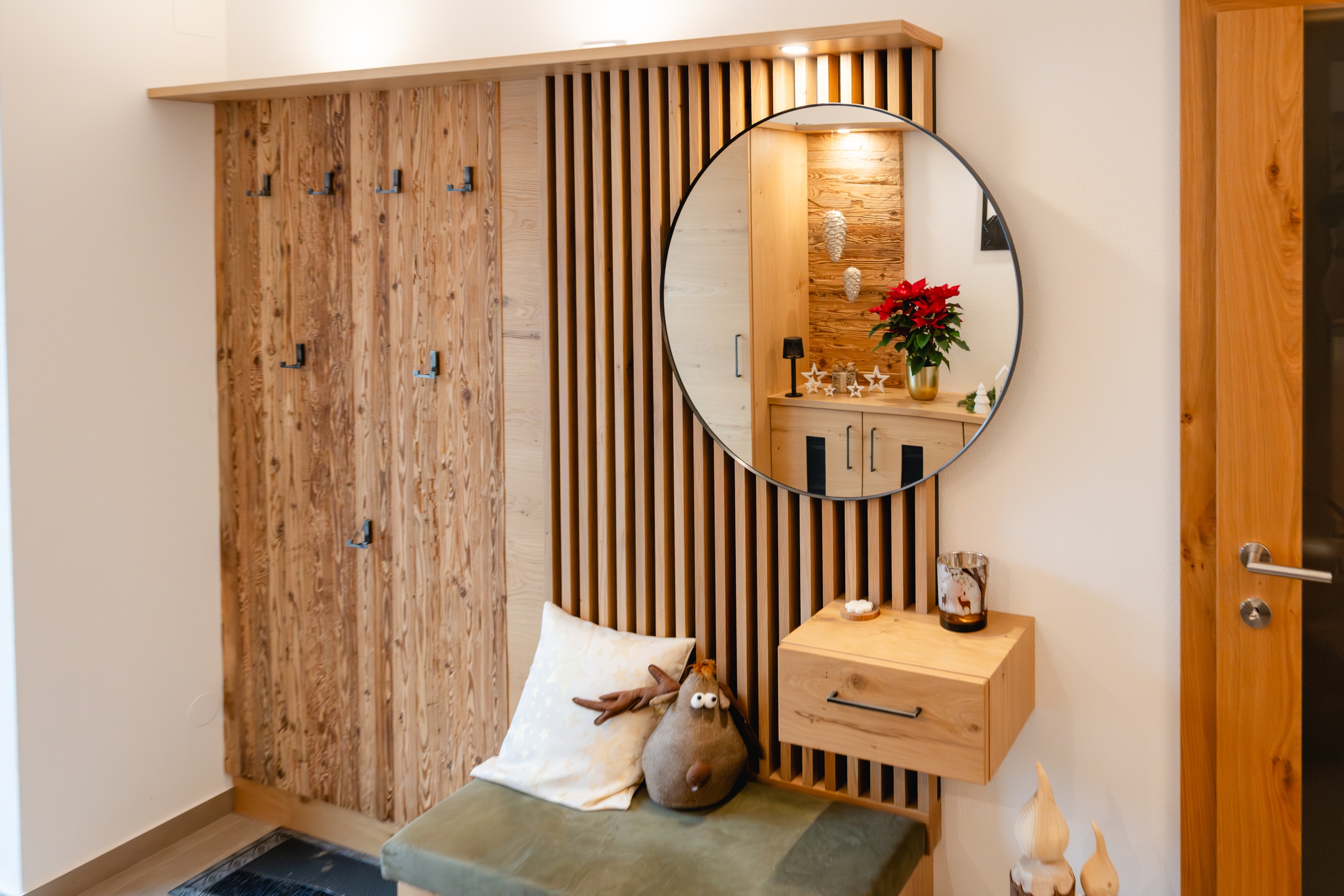 Ein Eingangs‑Garderobenbereich mit einer Rückwand im Stil-Mix aus Holzsprossen und stark verästeltem Holz, Spiegel, gepolsterter Sitzfläche und schwebenden Kästchen von Tischlerei Scharnreitner.