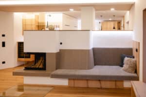 Ein Kamin mit gepolsterter grauer Sitzbank mit Lehne übergehend in eine elegante Keramik‑Lehne, harmonisch kombiniert mit warmen Holz‑Oberflächen von Tischlerei Scharnreitner.