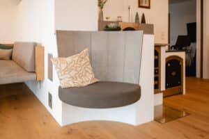 Ein Wohnzimmer mit angrenzendem Essbereich und Kamin mit gepolsterter runder Einzelsitzfläche, meisterlich gestaltet von Tischlerei Scharnreitner.