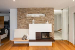 Ein Kamin mit integrierter Sitzfläche und Sitzbank vor einer Steinwand, kombiniert mit warmen Holz‑Akzenten in gemütlicher Atmosphäre von Tischlerei Scharnreitner.