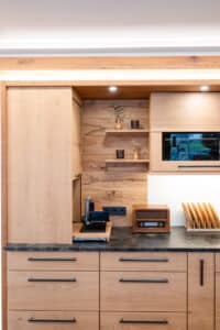 Eine Küchen mit Rolladenschrank, ausgestattet mit einem Auszug für Küchengeräte wie Allesschneider oder Brotmaschine, elegant integriert in warmer Holzoptik von Tischlerei Scharnreitner.