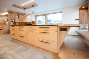 Ein offene Küche mit Kochinsel und Plattform‑Essbank in warmem Holzton, klaren Linien und durchdachtem Raumkonzept von Tischlerei Scharnreitner.